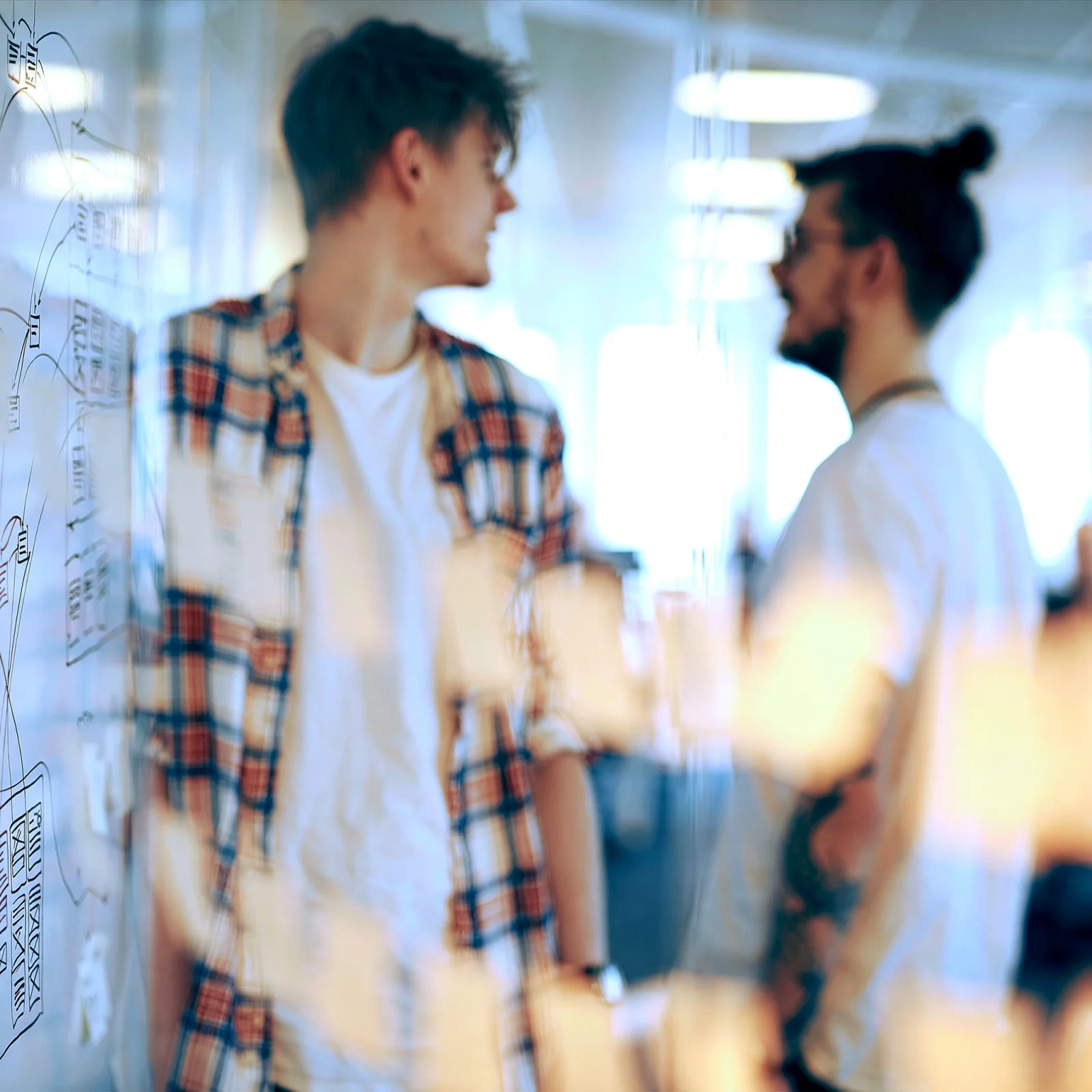 Two of the team talking at a glass planning board, shot wide open and blown out by window light